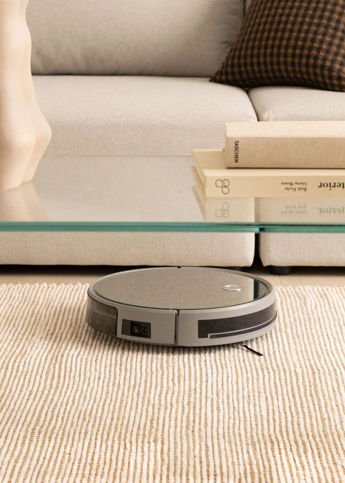 Aspiradora robot en alfombra rayada, con sofá y libros al fondo.