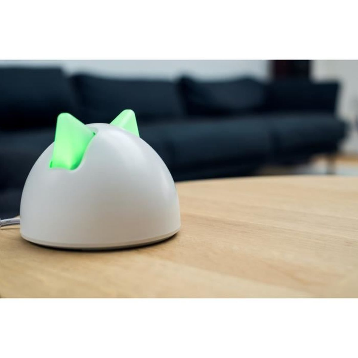 Weißes, katzenförmiges Gerät mit grünen Lichtern auf einem Holztisch. Schwarzes Sofa im Hintergrund.