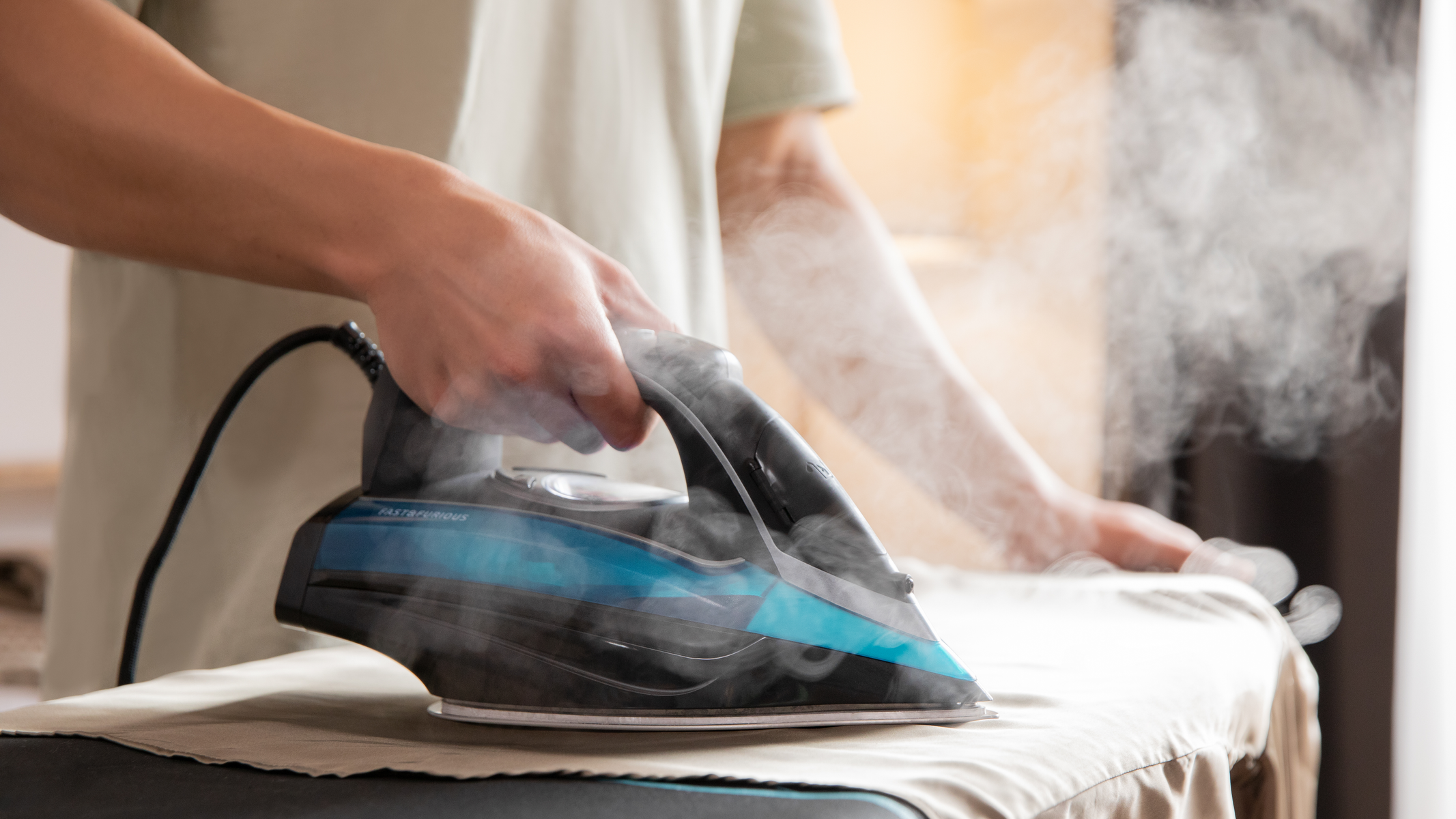 Osoba prasująca ubrania. Para unosi się z żelazka. Żelazko jest niebiesko-czarne, na desce do prasowania.