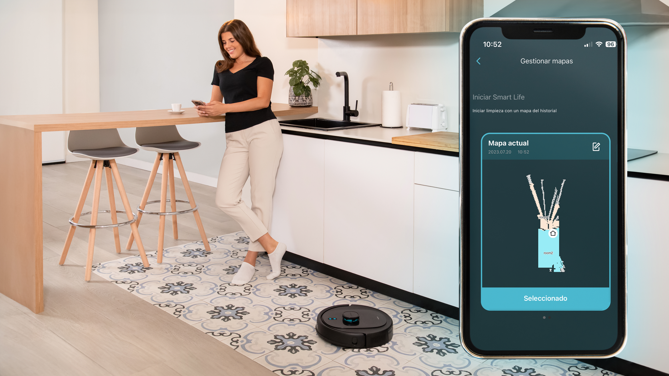 Mujer en la cocina con teléfono y un robot aspirador. El teléfono muestra la interfaz de la app de mapa de limpieza.