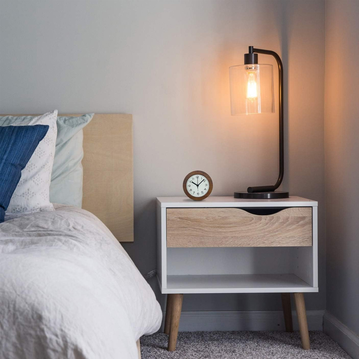 Nachttisch mit Lampe, Uhr und Schublade neben einem Bett.