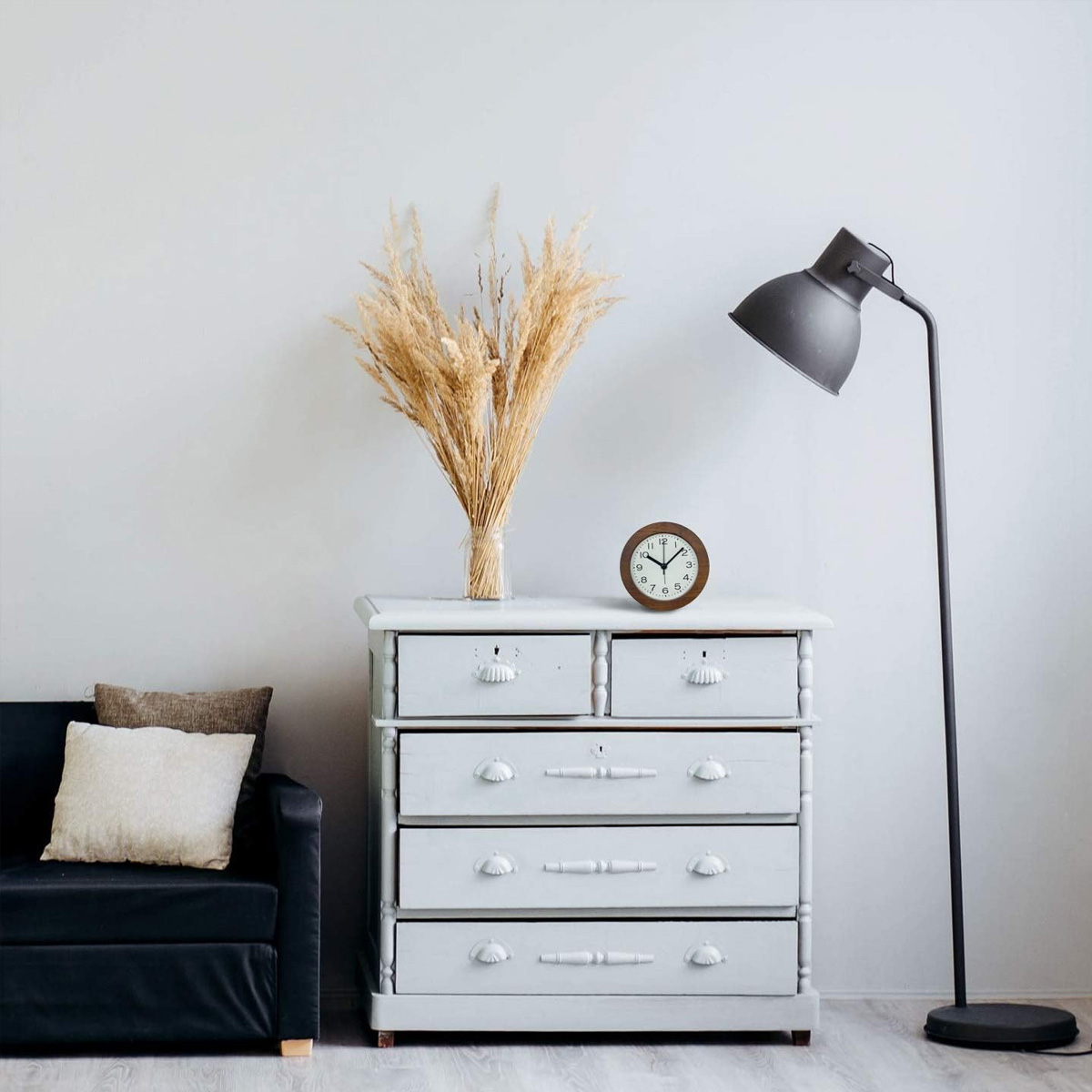 Ein Raum mit einer weißen Kommode, getrockneten Pflanzen, einer Uhr, einer schwarzen Couch und einer Stehlampe.