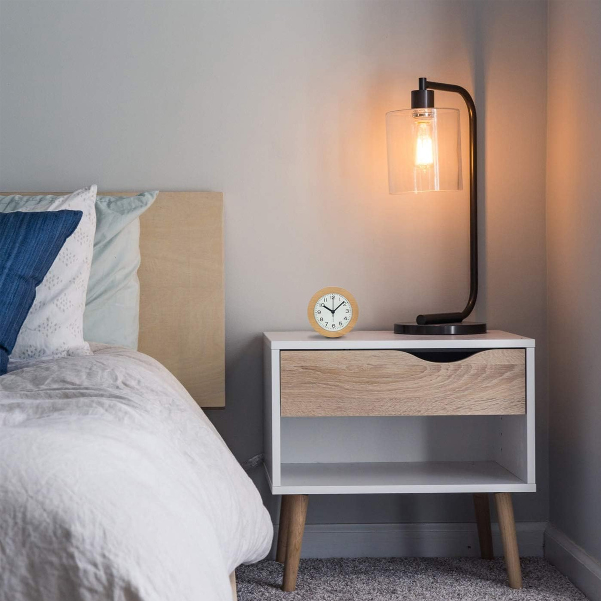 Nachttisch mit Lampe und Wecker. Bett und Kissen links. Weiche Beleuchtung.