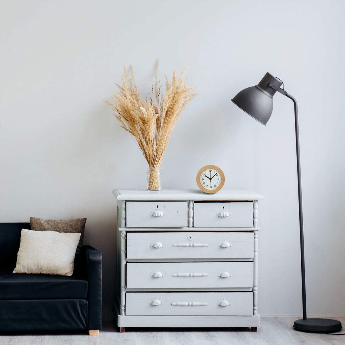 Eine Zimmerecke mit Kommode, Lampe, schwarzem Sofa und getrockneten Pflanzen in einer Vase.