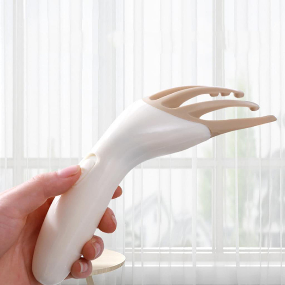 Eine Hand hält ein weiß-beige Kopfhautmassagegerät. Im Hintergrund ist ein Fenster mit weißen Vorhängen zu sehen.