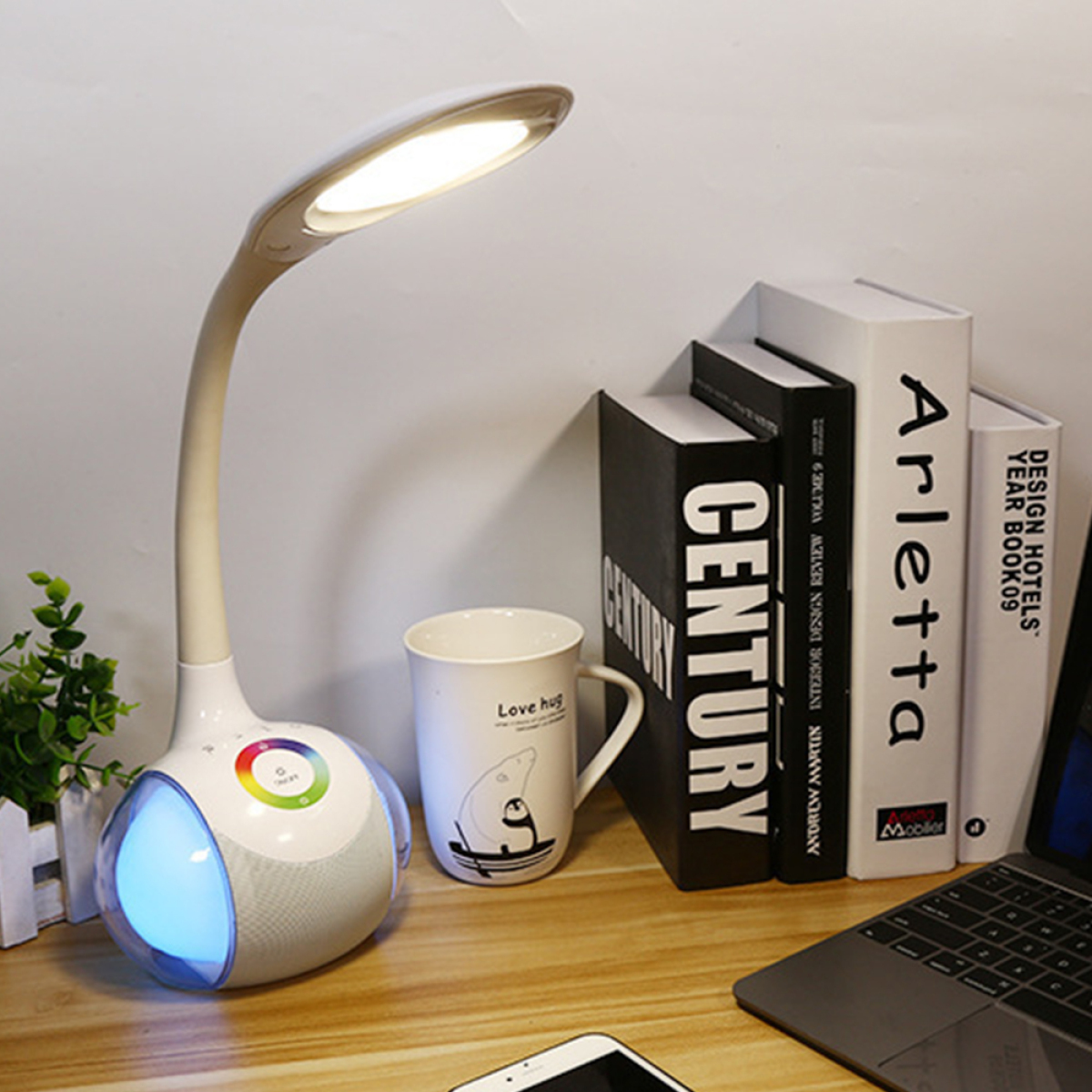 Schreibtischlampe mit Lautsprecher, Büchern, Becher und Laptop. Holzschreibtisch, weiße Wand im Hintergrund.