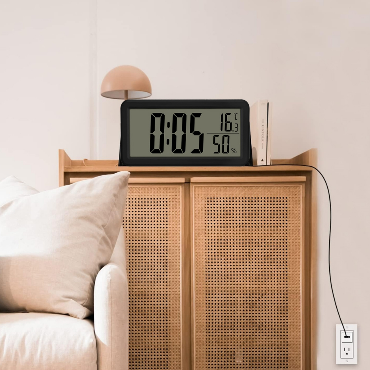 Eine Digitaluhr zeigt Zeit, Temperatur und Luftfeuchtigkeit an. Sie steht auf einem Holzschrank mit Lampe und Büchern.