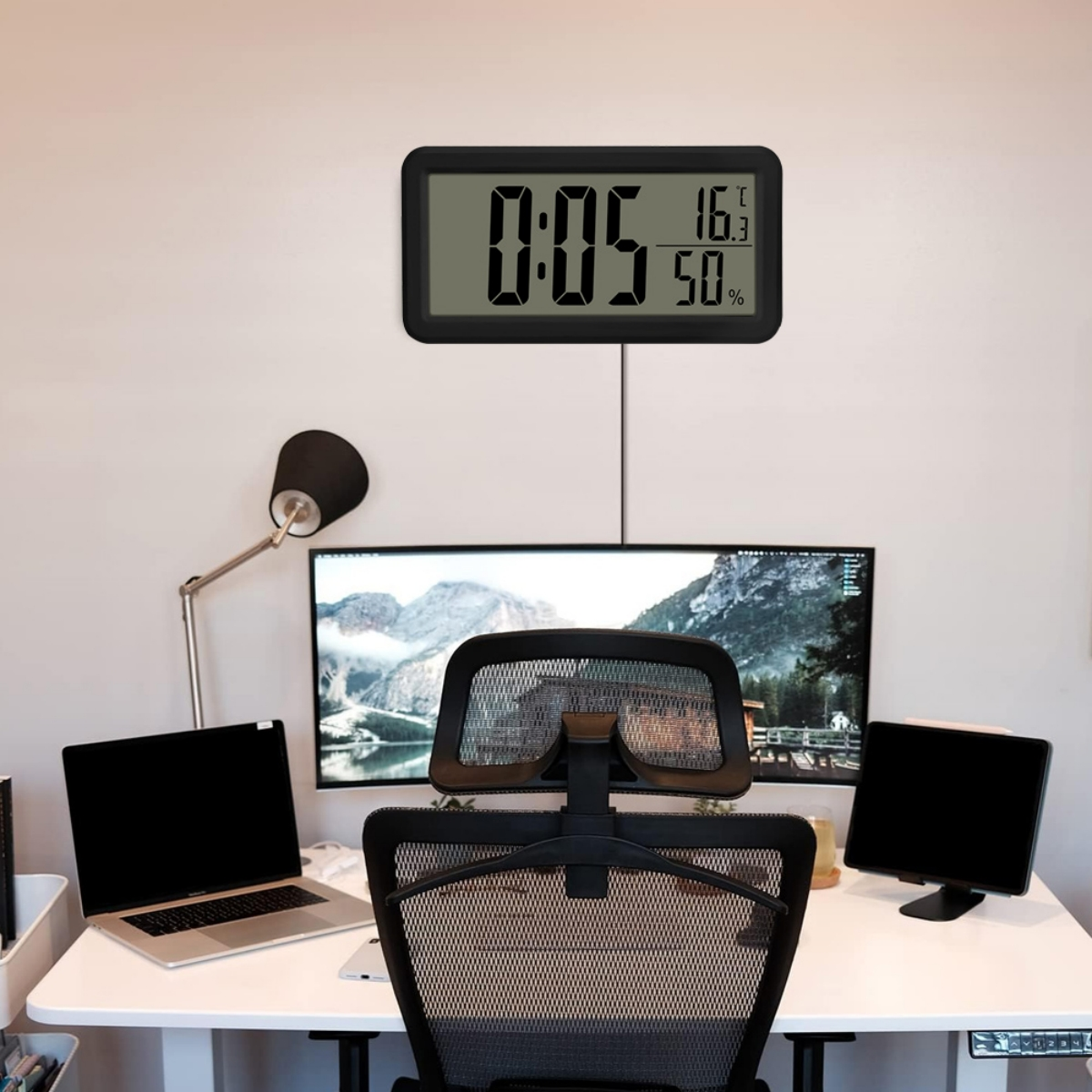 Eine Digitaluhr zeigt Zeit, Temperatur und Luftfeuchtigkeit an einer Wand über einem Schreibtisch mit Monitoren.