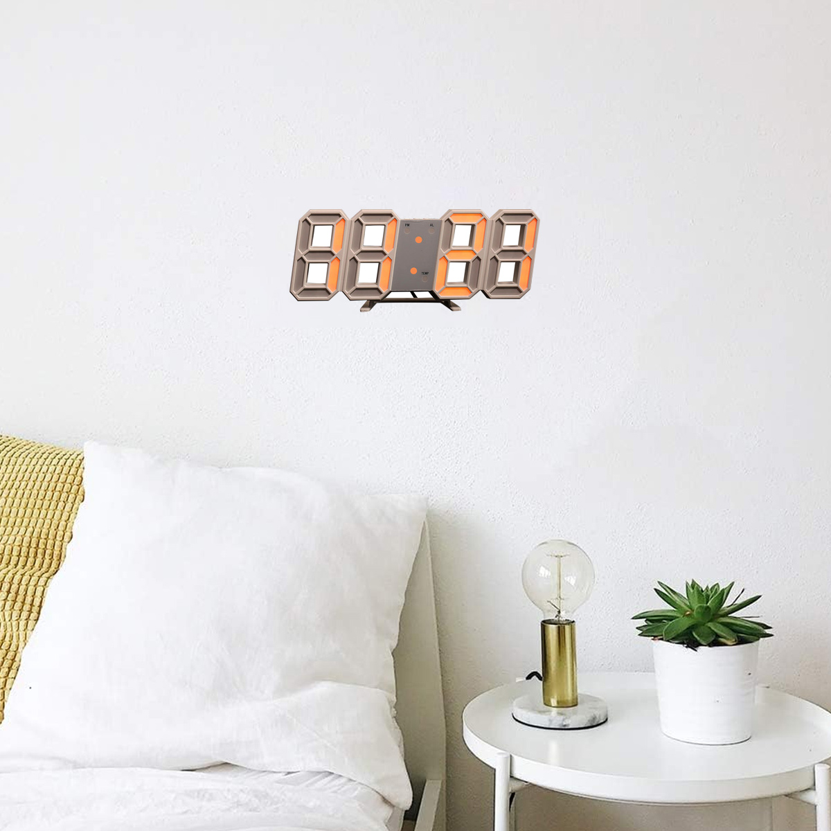 Eine Digitaluhr an einer weißen Wand. Eine kleine Pflanze und eine Lampe stehen auf einem Tisch unter der Uhr.