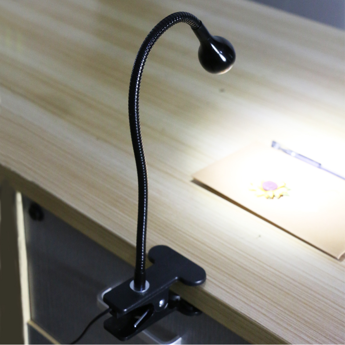 Eine schwarze Schreibtischlampe mit flexiblem Hals, an einer Holzoberfläche befestigt. Die Lampe leuchtet.