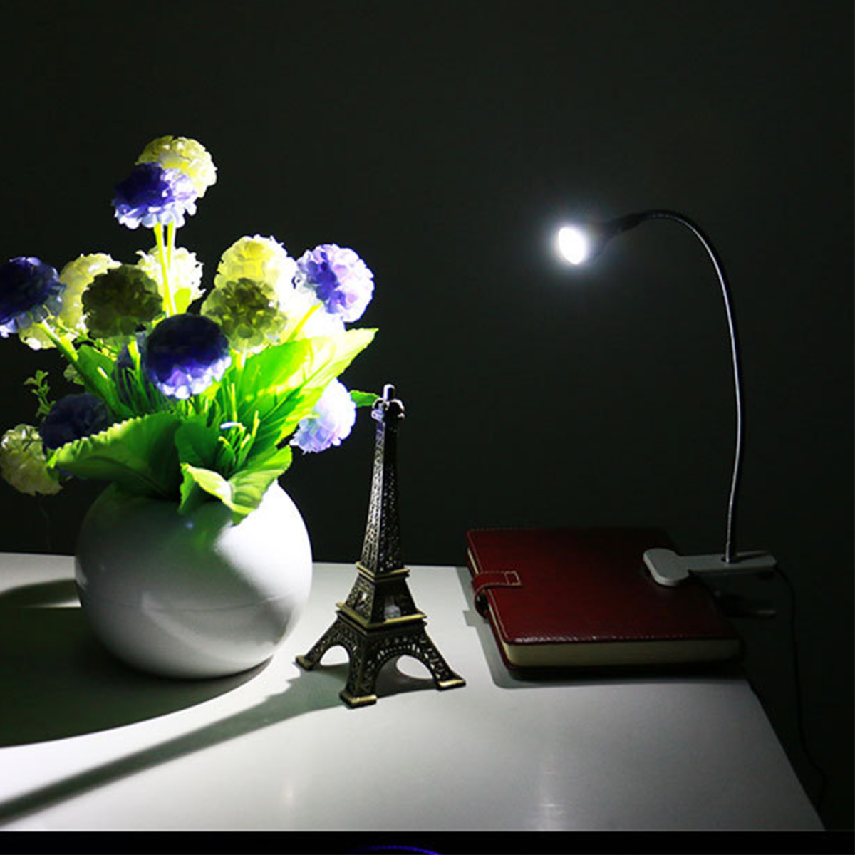 Ein Schreibtisch mit Blumenvase, Eiffelturm, Notizbuch und kleiner Lampe. Dunkler Hintergrund.