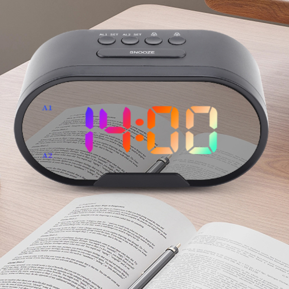 Schwarzer digitaler Wecker mit verspiegelter Oberfläche, die 14:00 anzeigt. Tasten für Einstellungen und Schlummerfunktion. Offenes Buch sichtbar.