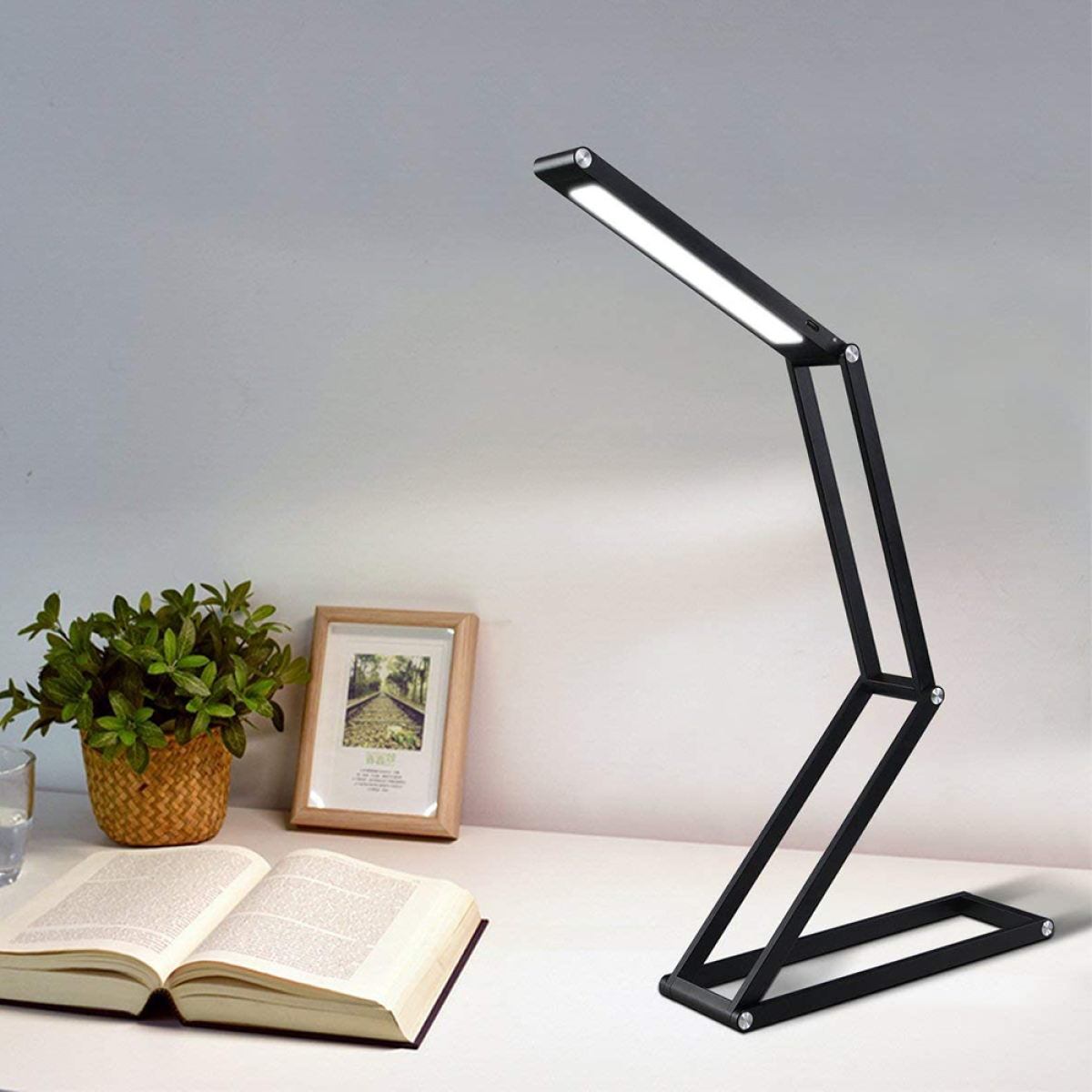 Schwarze, zusammenklappbare Schreibtischlampe auf weißem Schreibtisch mit Pflanze und Buch.