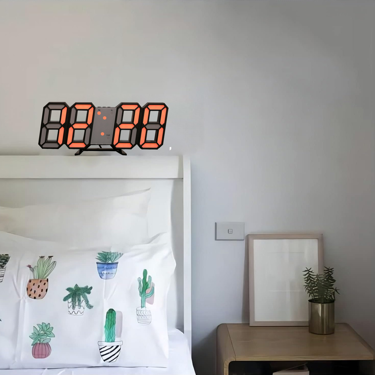 Digitaluhr an einer Wand über einem Bett. Kissen mit Pflanzendesigns, Rahmen und kleiner Pflanze.
