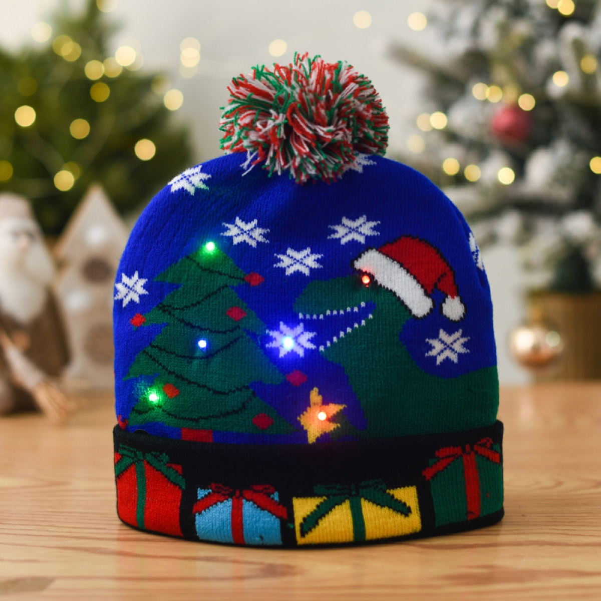 Eine blaue Weihnachtsmütze mit Dinosaurier, Weihnachtsbaum und Geschenken, mit bunten Lichtern und Bommel.
