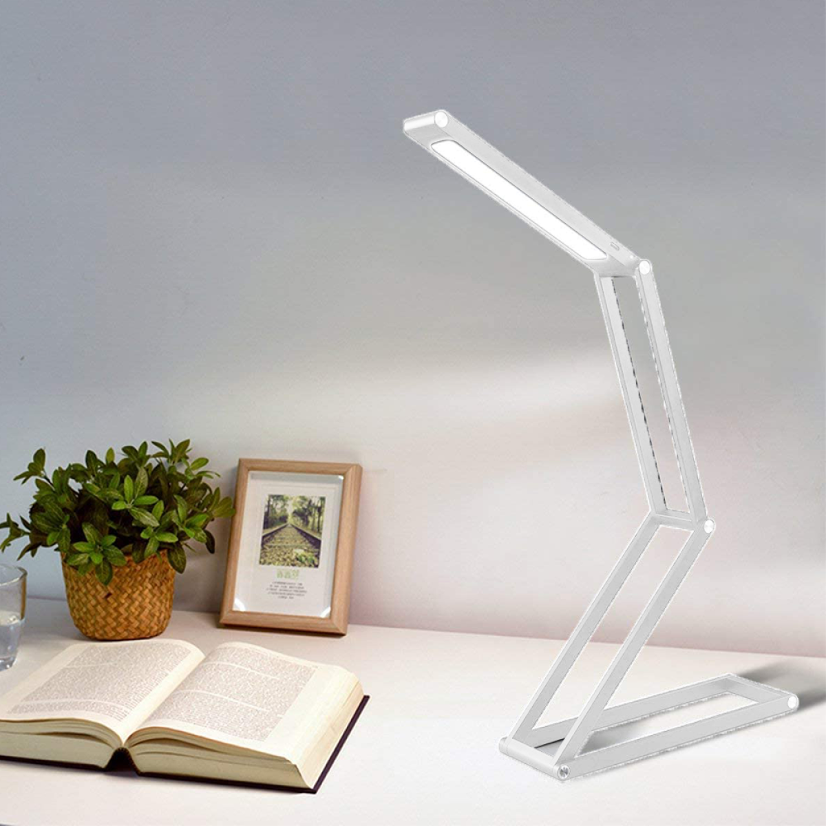 Eine weiße, faltbare Schreibtischlampe steht auf einem weißen Schreibtisch, im Hintergrund ein Buch und eine Pflanze.