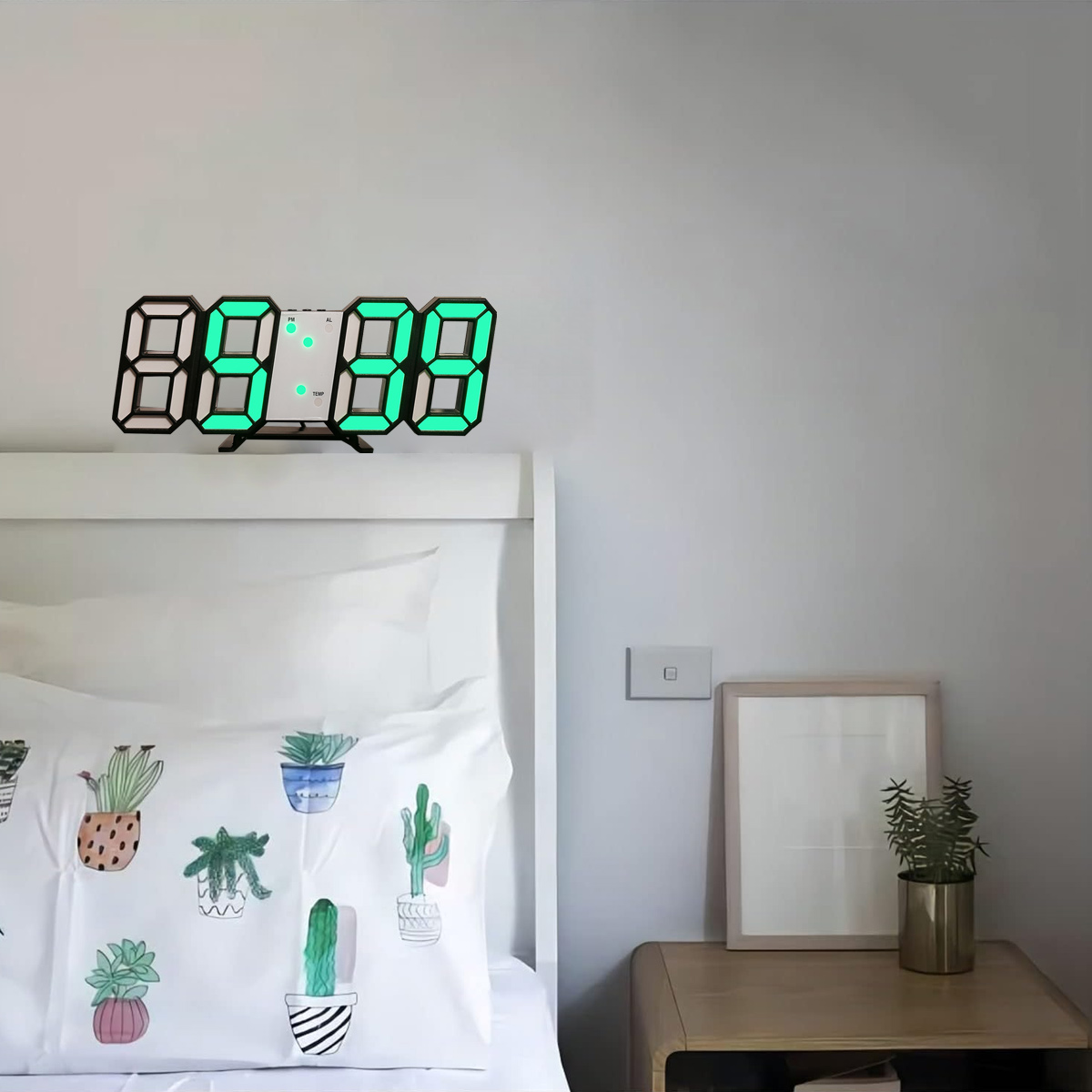 Digitaluhr mit grünen Ziffern, an weißer Wand über Bett montiert. Kaktuskissen und Nachttisch aus Holz.