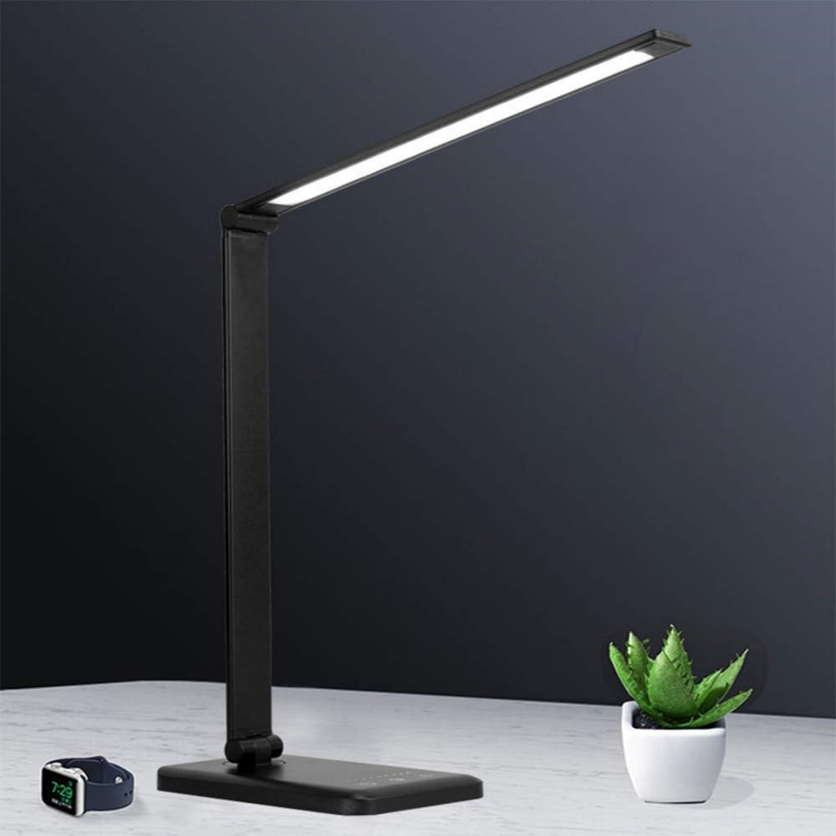 Schwarze Schreibtischlampe auf einem Tisch mit kleiner Pflanze und Smartwatch, dunkler Hintergrund.