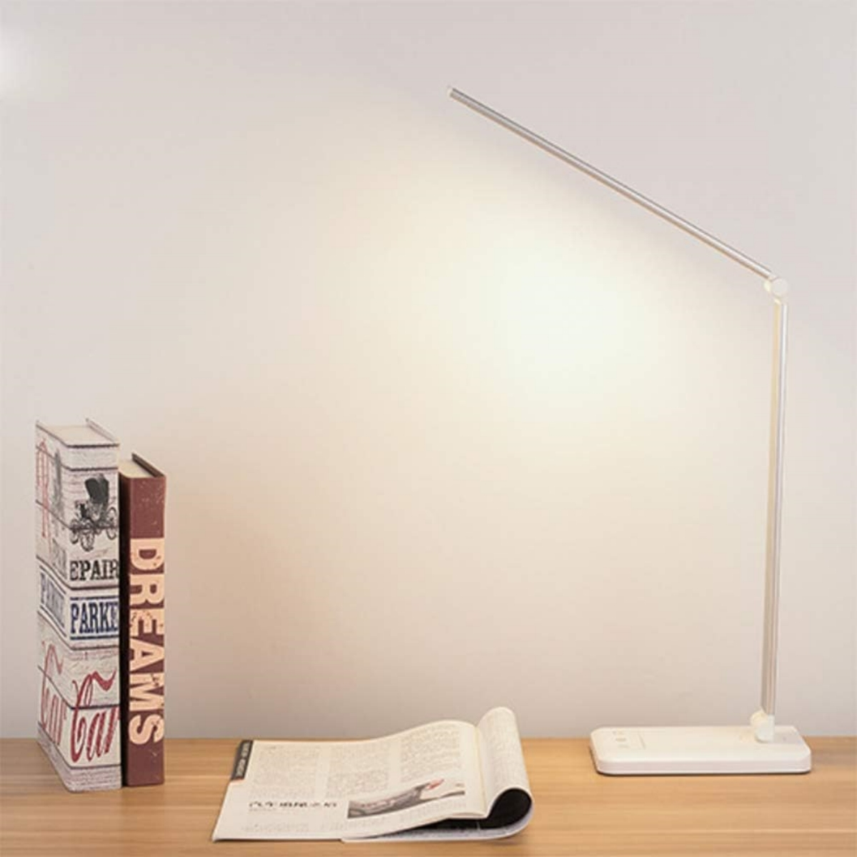 Eine moderne silberne Schreibtischlampe ist beleuchtet. Bücher und eine offene Zeitschrift liegen auf einem Holztisch.