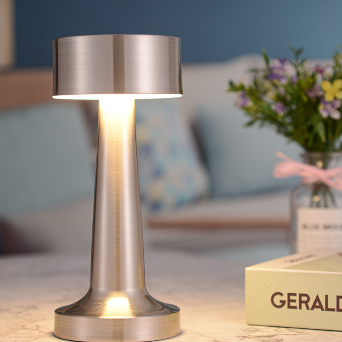 Metalltischlampe mit hellem Licht. Blumen und ein Buch stehen auf dem Tisch, mit blauem Wandhintergrund.