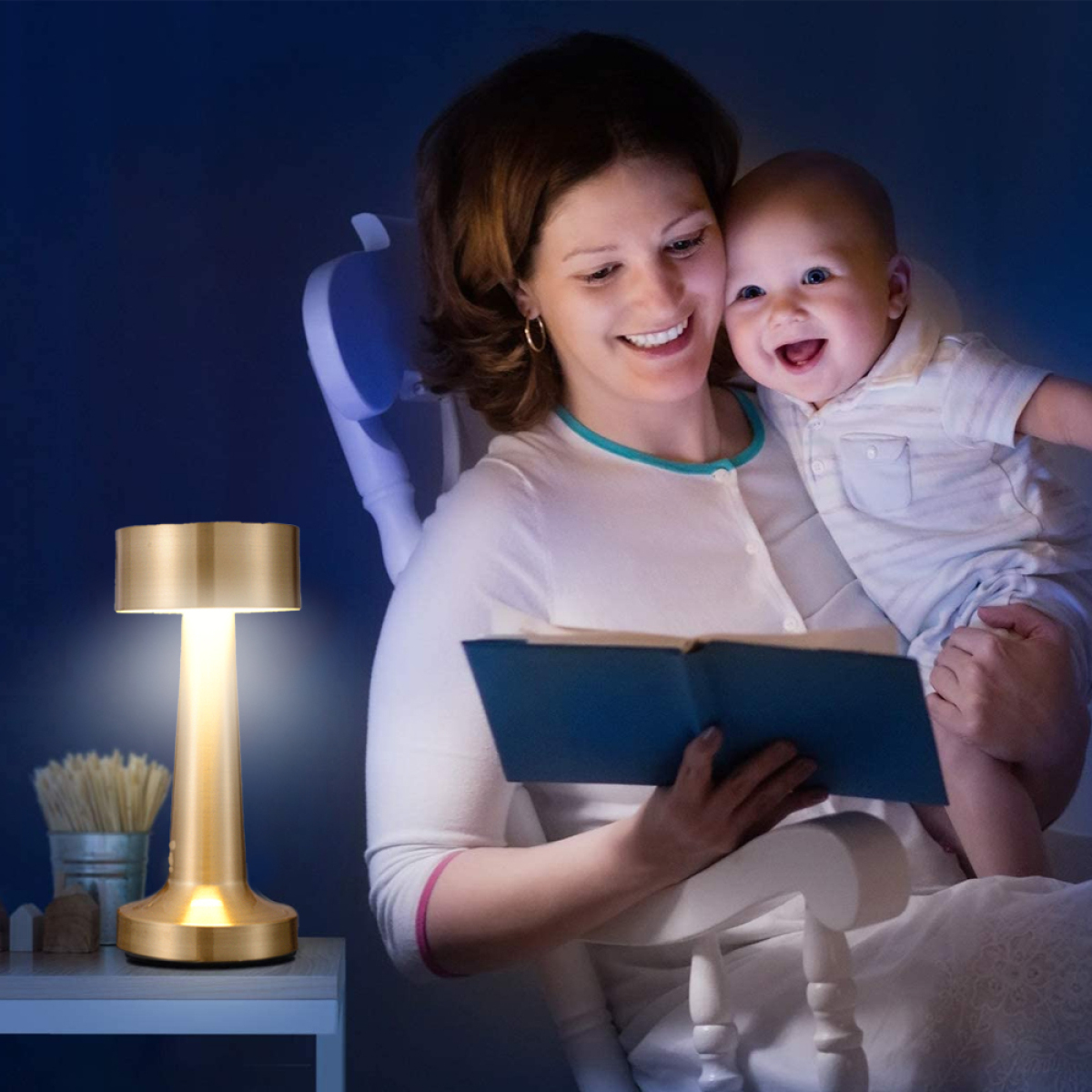 Frau hält Baby, liest Buch unter goldener Lampe. Baby lächelt. Weißer Stuhl, kleiner Tisch.