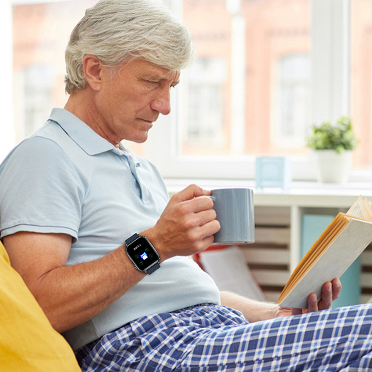 Älterer Mann liest Buch, hält Tasse, trägt Smartwatch. Sitzt auf Couch.