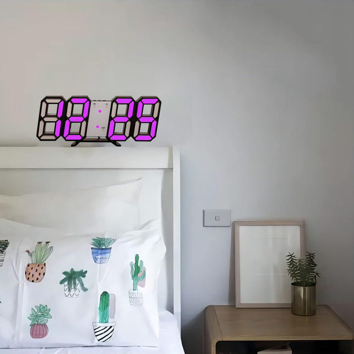 Digitaluhr mit 8:28 Uhr, auf einem weißen Bett mit einem Kaktuskissen und einem Bilderrahmen.