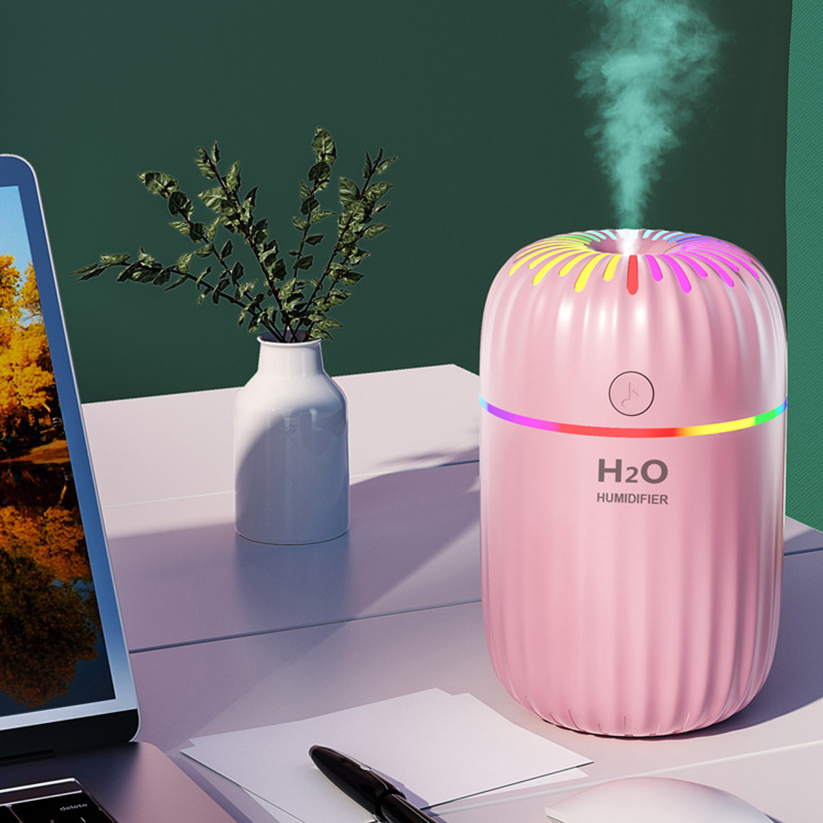 Ein rosa Luftbefeuchter mit farbenfroher Oberseite. Er steht auf einem weißen Schreibtisch mit einer Vase und einem Laptop.