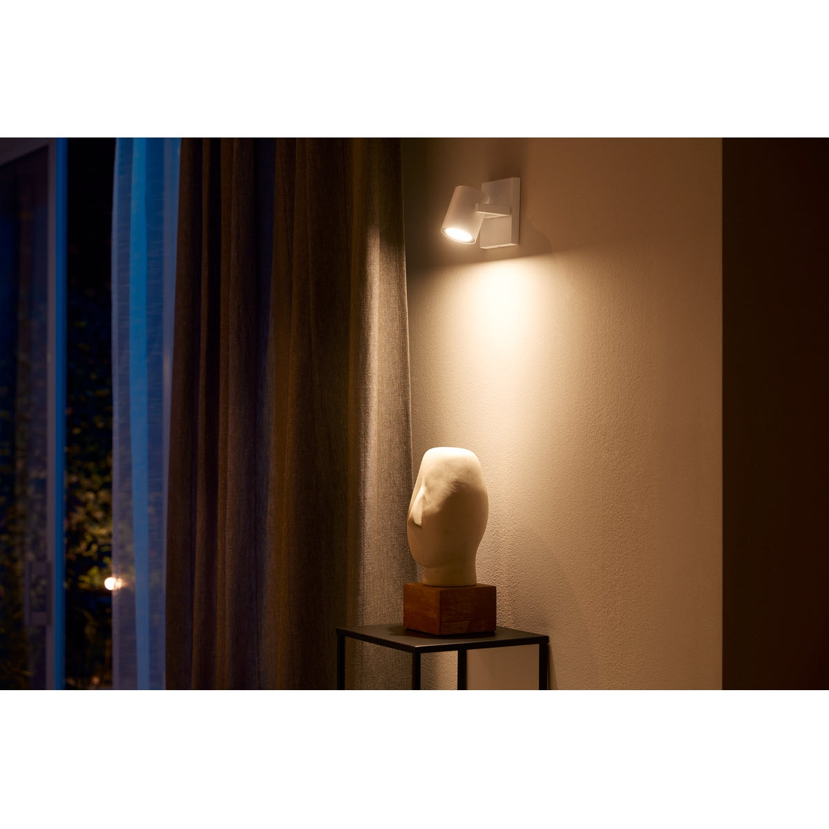 Una scultura di una testa su un supporto, illuminata da due luci a parete. Una tenda scura è sullo sfondo.