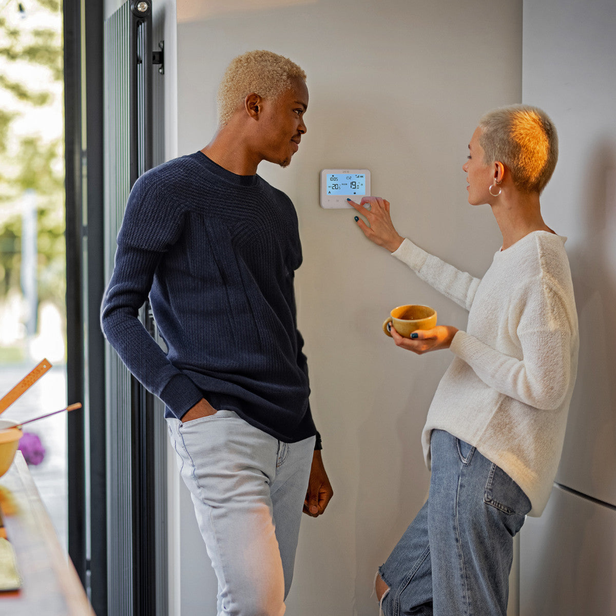 Una pareja está junto a una pared con un termostato. Uno ajusta la temperatura, el otro sostiene una taza.