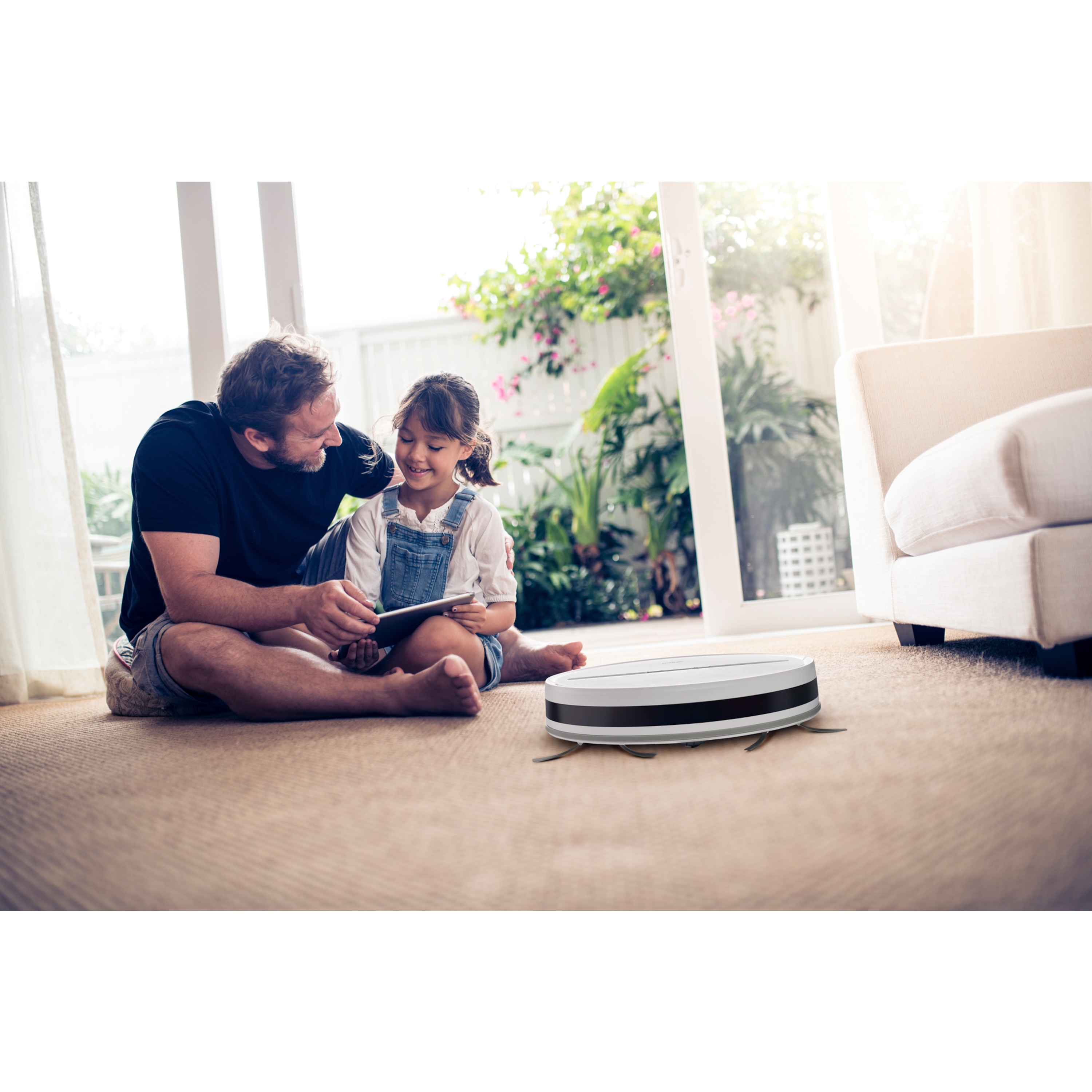 Ojciec i córka siedzą na dywanie. Odkurzacz robot znajduje się na podłodze w pobliżu.