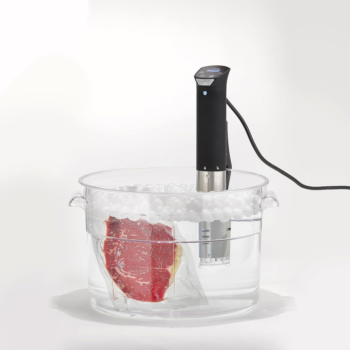 Sous-Vide-Gerät in einem transparenten Behälter mit Steak in einem versiegelten Beutel. Wasser und weiße Kugeln sind vorhanden.