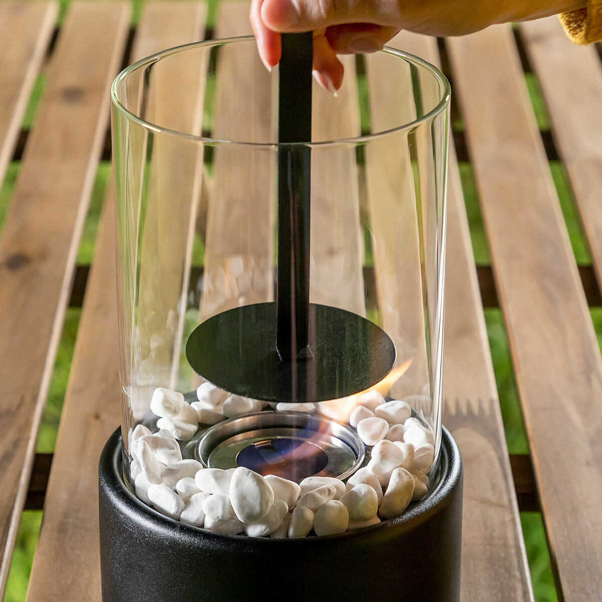 Glas-Feuerstelle mit weißen Steinen und Flamme. Schwarzer Sockel, Hand hält Werkzeug über Flamme.