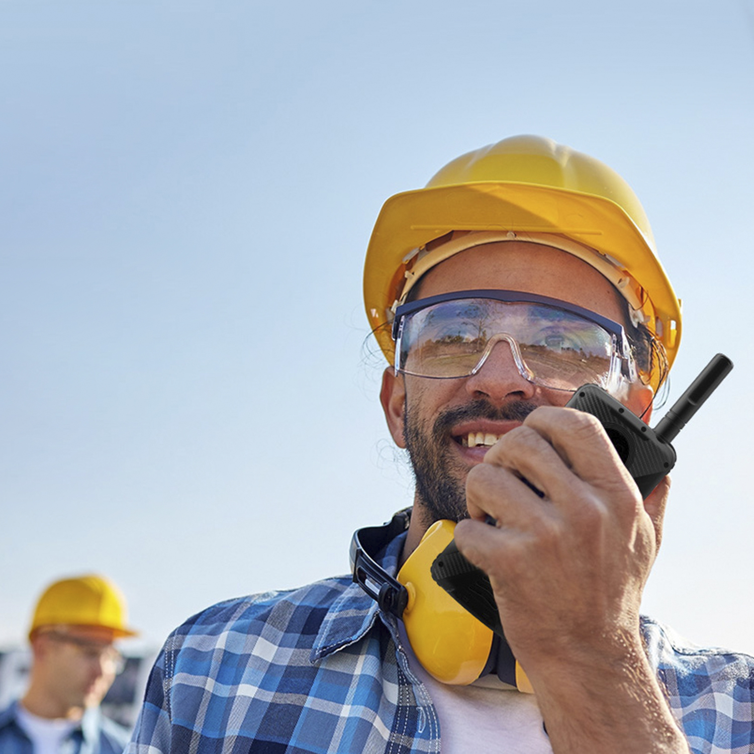 Un operaio edile con casco giallo e occhiali protettivi, che tiene un walkie-talkie. Un altro lavoratore sullo sfondo.