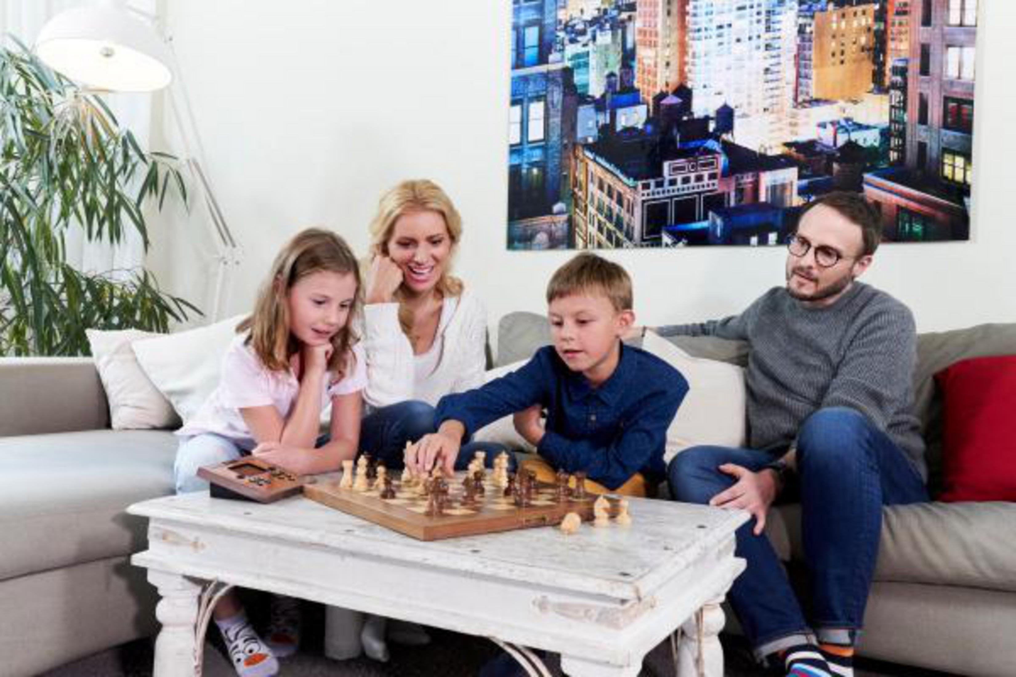 Una famiglia gioca a scacchi a un tavolo. Un dipinto di città è appeso al muro sullo sfondo.