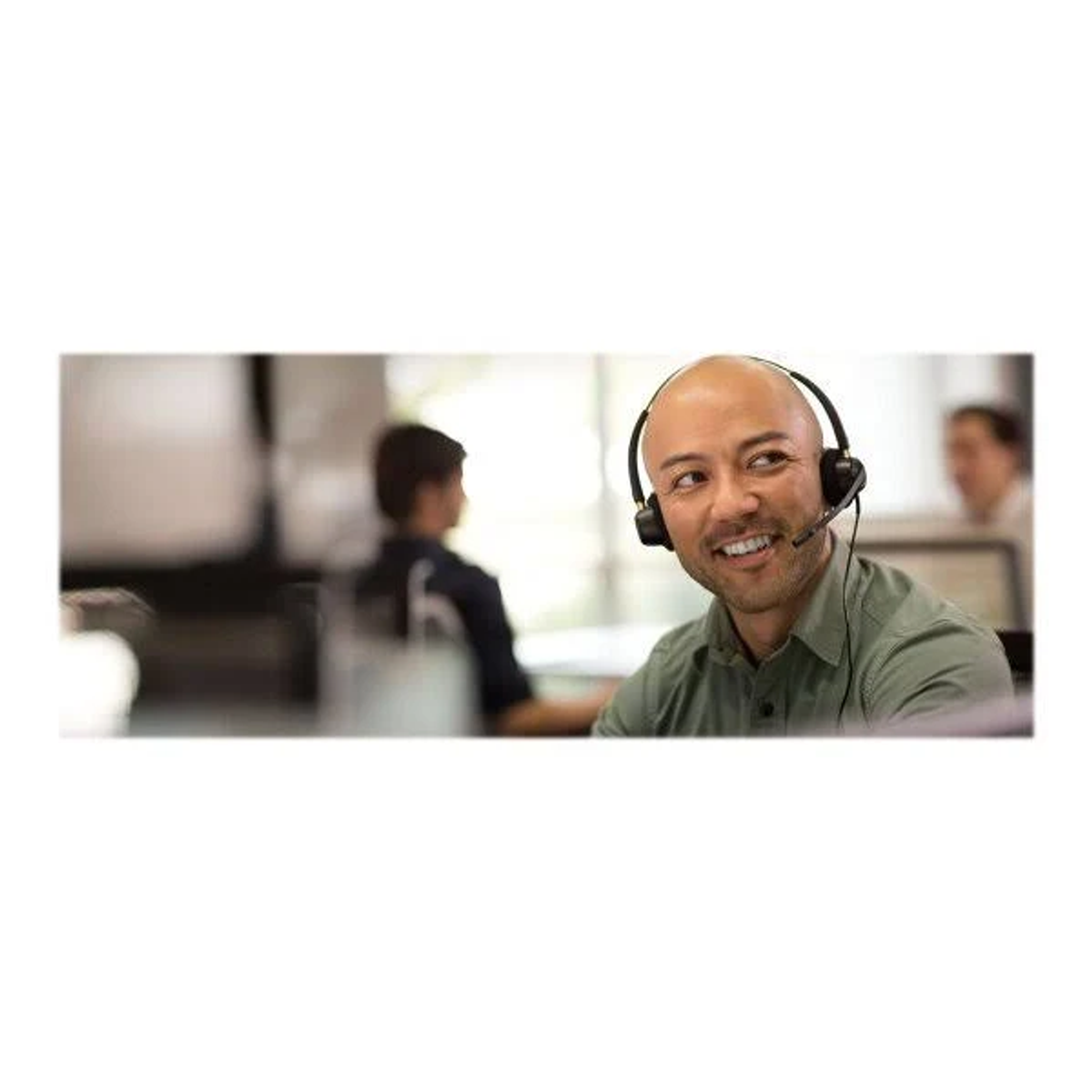 Smiling man with headset in an office setting, with colleagues blurred in the background.