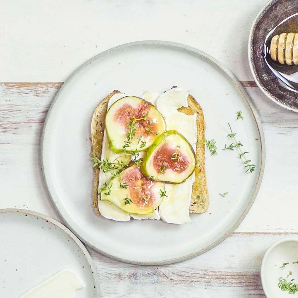 Toast mit Brie, Feigen und Thymian auf einem weißen Teller. Andere Teller und Zutaten sind drum herum.