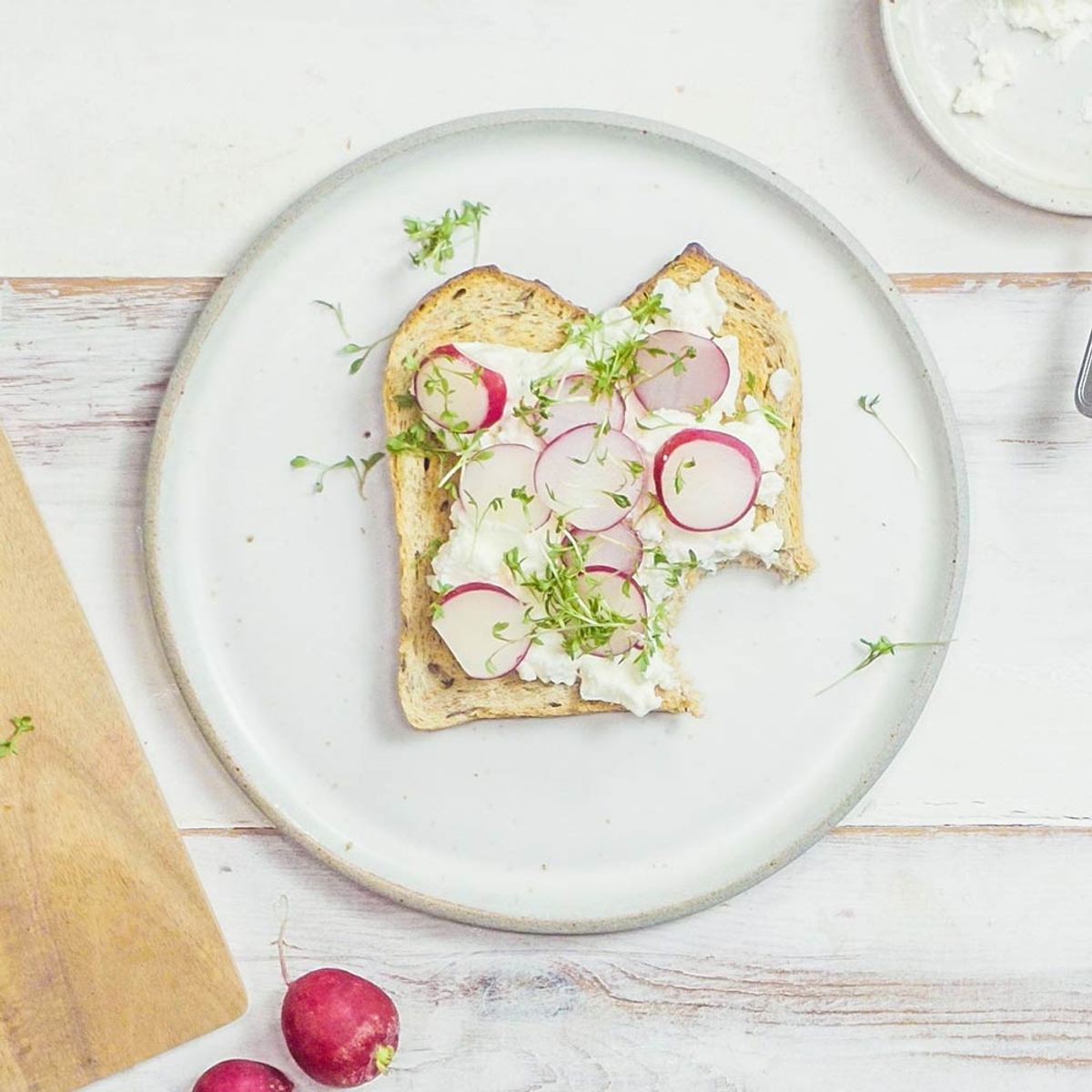 Toast mit Frischkäse, Radieschen und Microgreens auf einem Teller. Holzschneidebrett links.