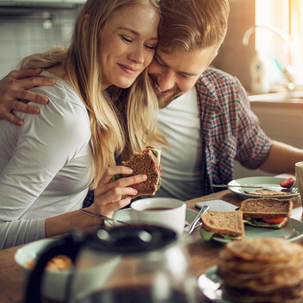Ein lächelndes Paar, das Sandwiches an einem Tisch isst, mit einer Kaffeekanne in der Nähe.