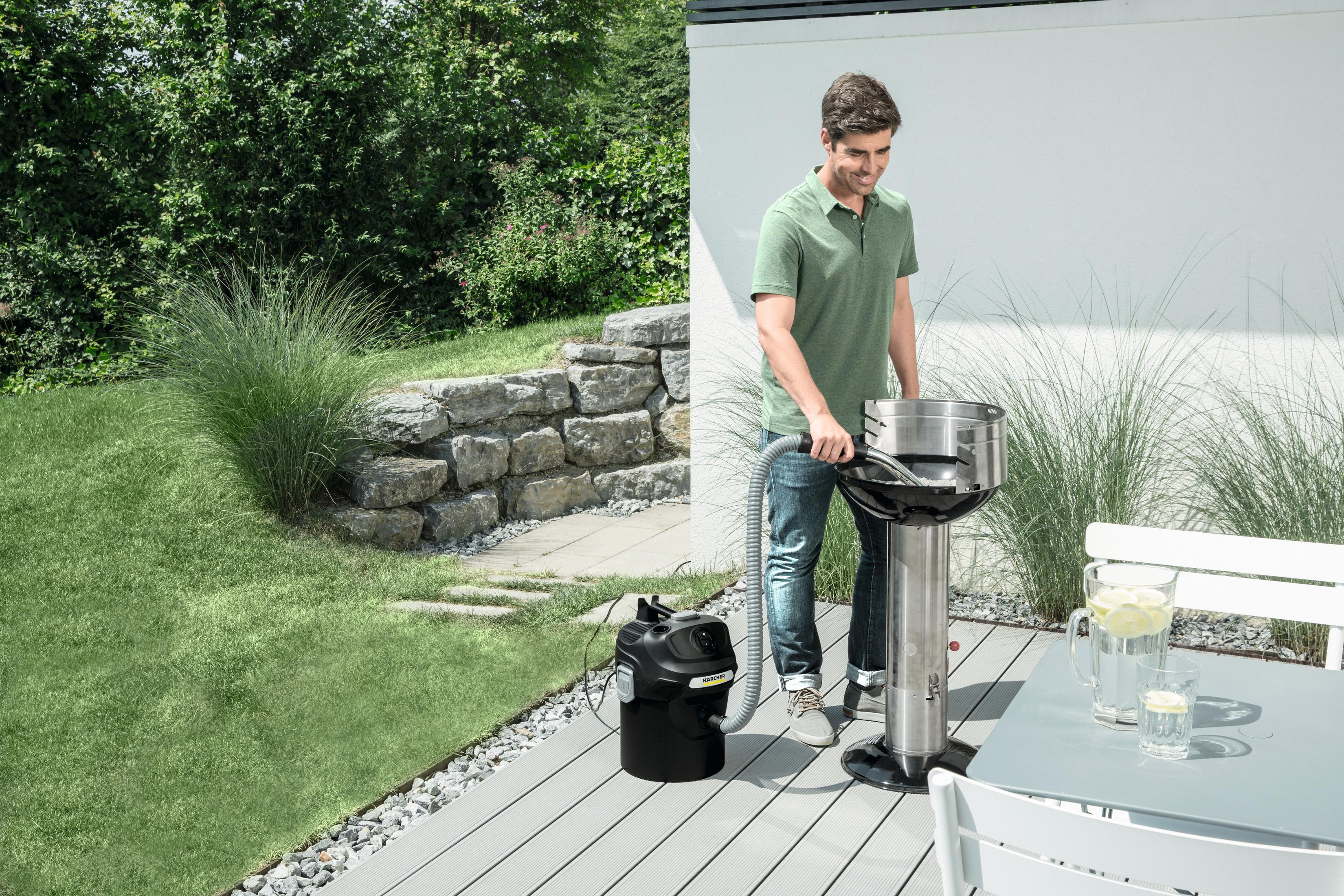 Mężczyzna odkurza grilla. Po lewej stronie znajduje się czarny odkurzacz. Widoczna jest zielona trawa, patio i stół.