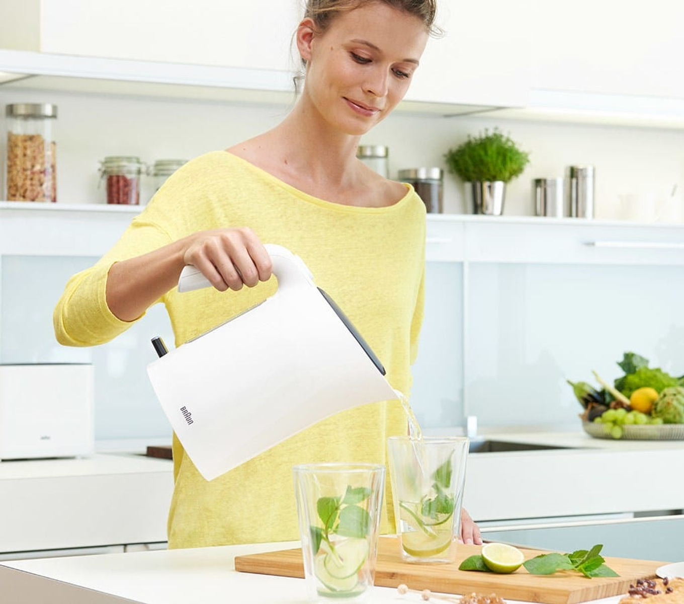 Kobieta w kuchni nalewa wodę z białego czajnika do dwóch szklanek z plasterkami limonki i miętą.