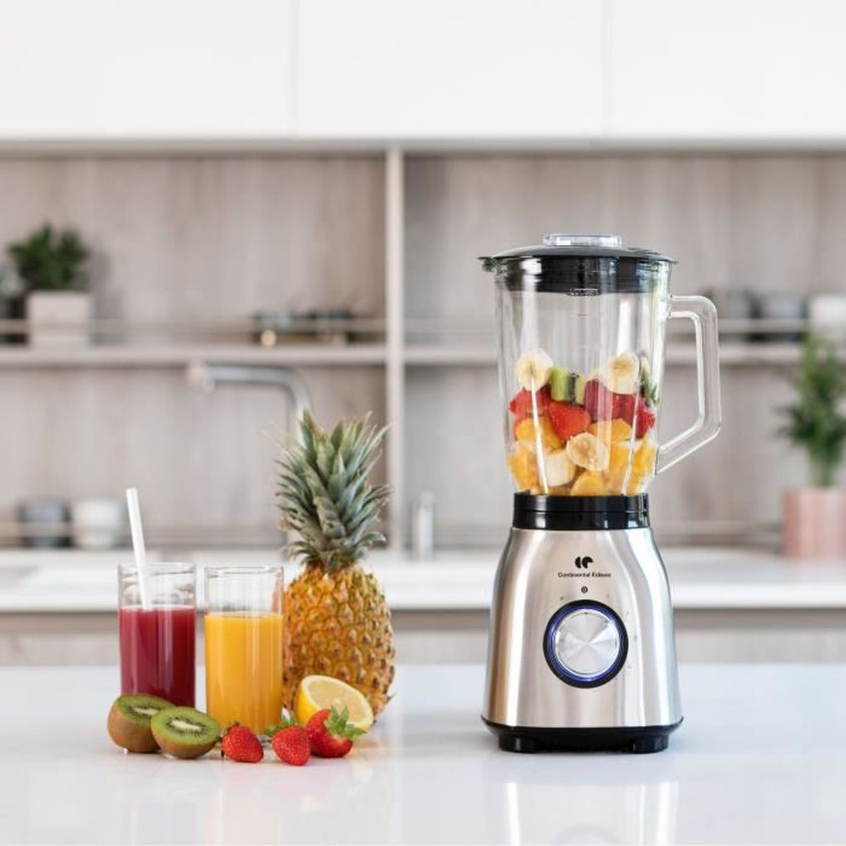 Un frullatore con frutta all'interno, due bicchieri di succo, e un ananas e kiwi su una superficie bianca.