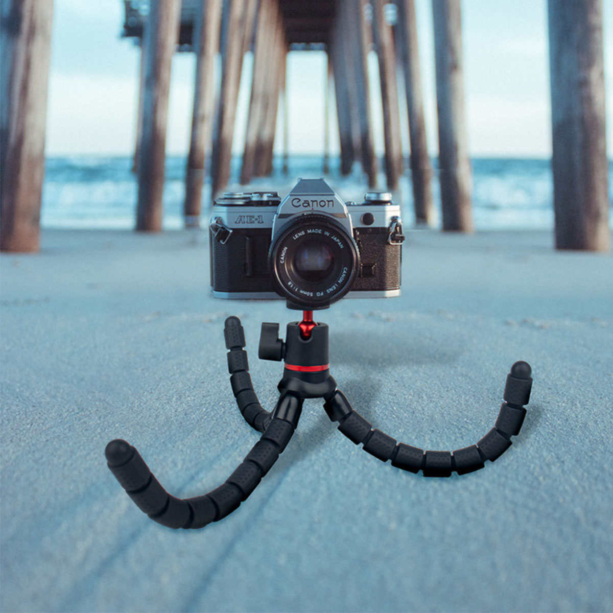 Eine Canon-Kamera auf einem flexiblen Stativ an einem Sandstrand, unter einem Pier.