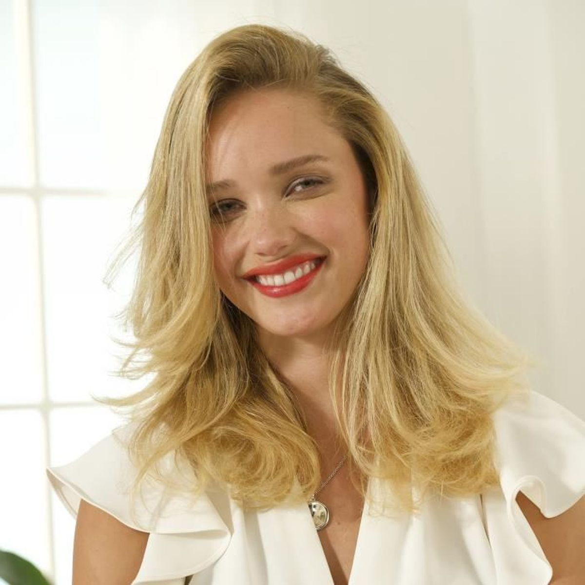 Blondynka z czerwoną szminką uśmiecha się. Ma długie włosy i białą bluzkę.