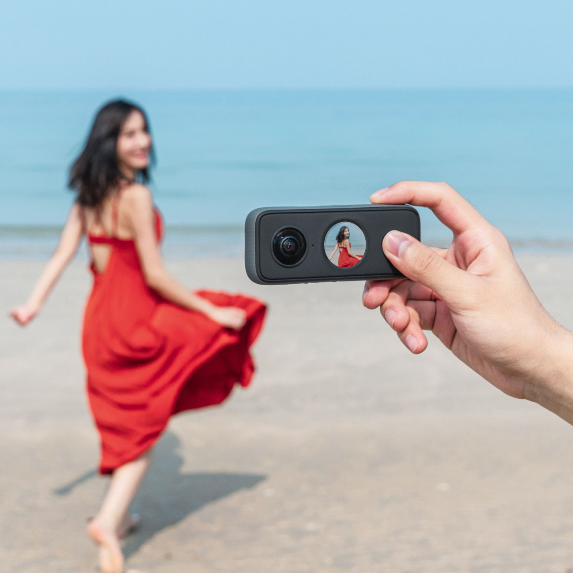 Eine Person hält eine Kamera, die eine Frau in rotem Kleid am Strand am Meer zeigt.