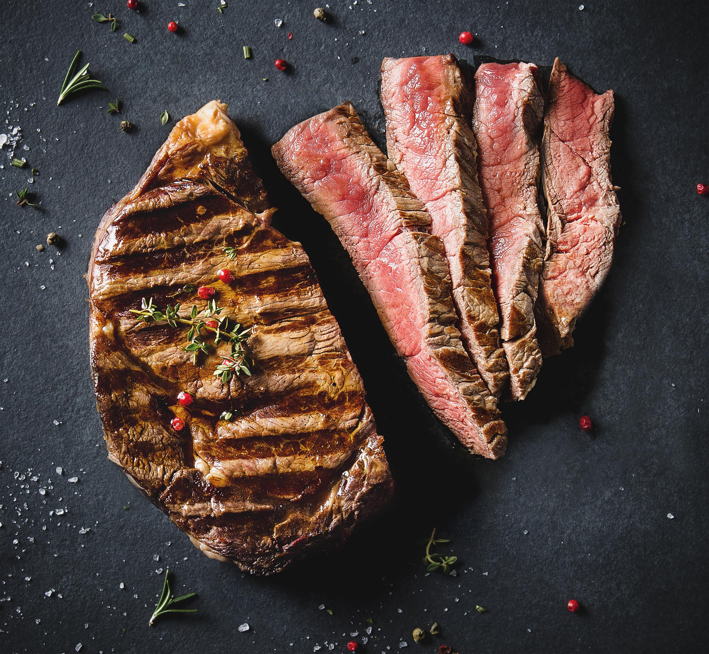 Grillowany stek, pokrojony i w całości. Ozdobiony rozmarynem i ziarnami pieprzu na ciemnej powierzchni.