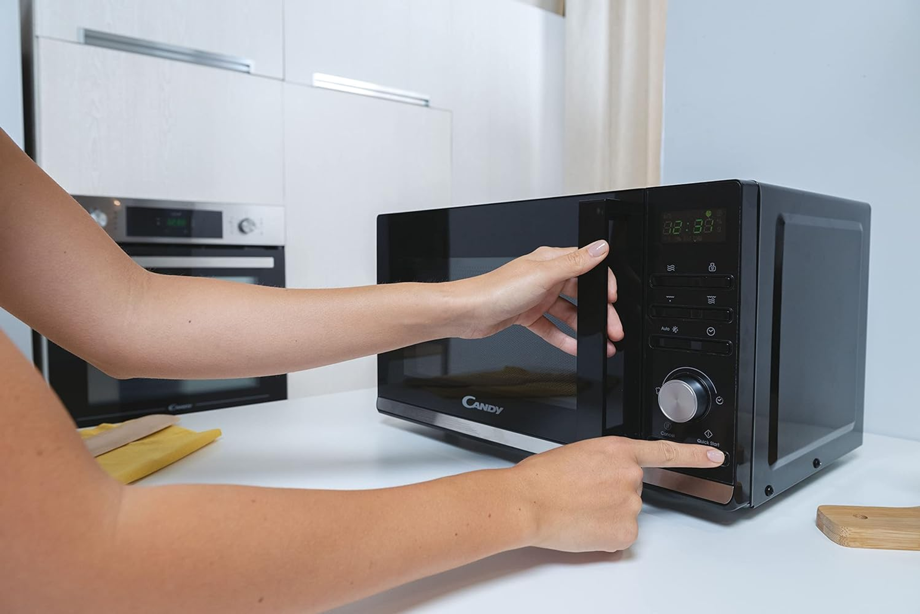 Ręce kobiety otwierające czarną kuchenkę mikrofalową na białym blacie w kuchni.