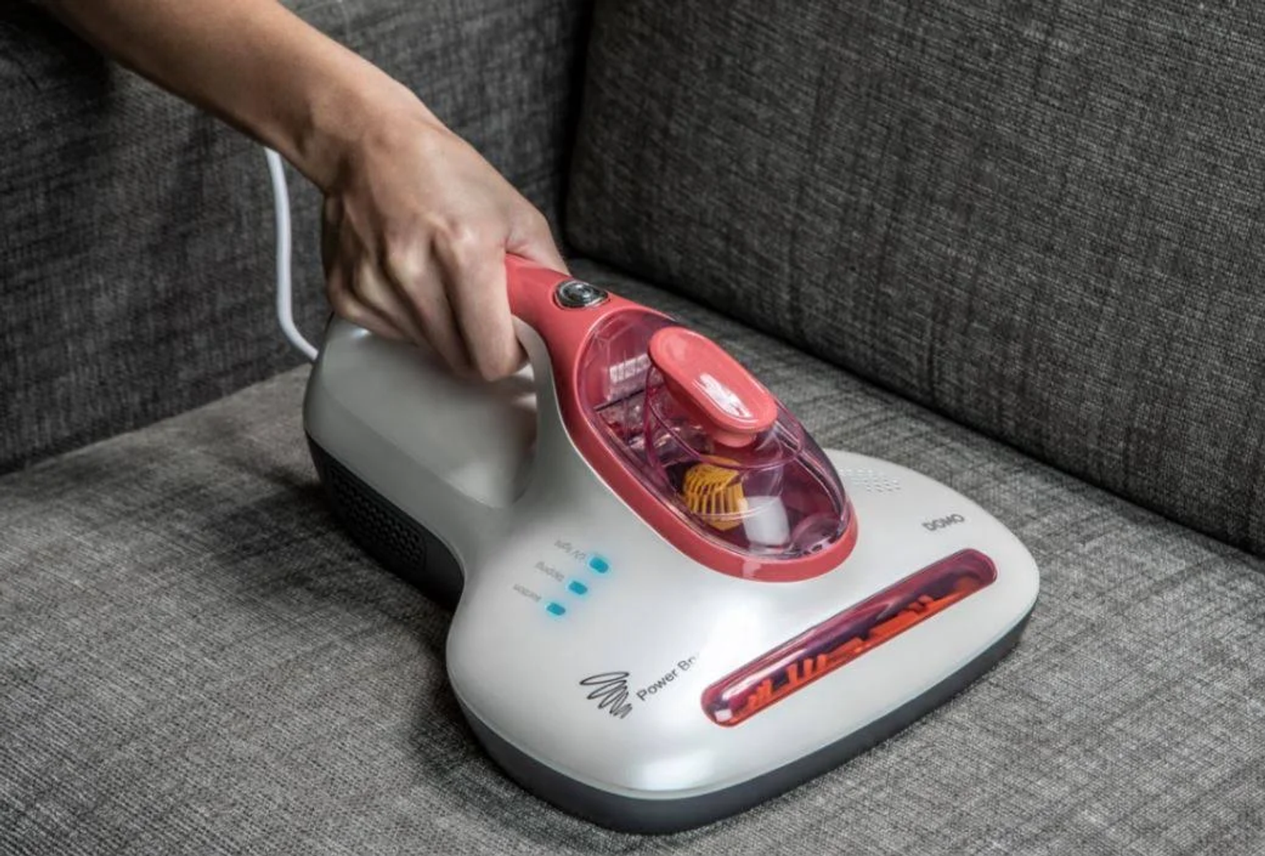 Una mano che tiene un aspirapolvere portatile DOMO bianco e rosso su un divano grigio. La spazzola elettrica è evidenziata.