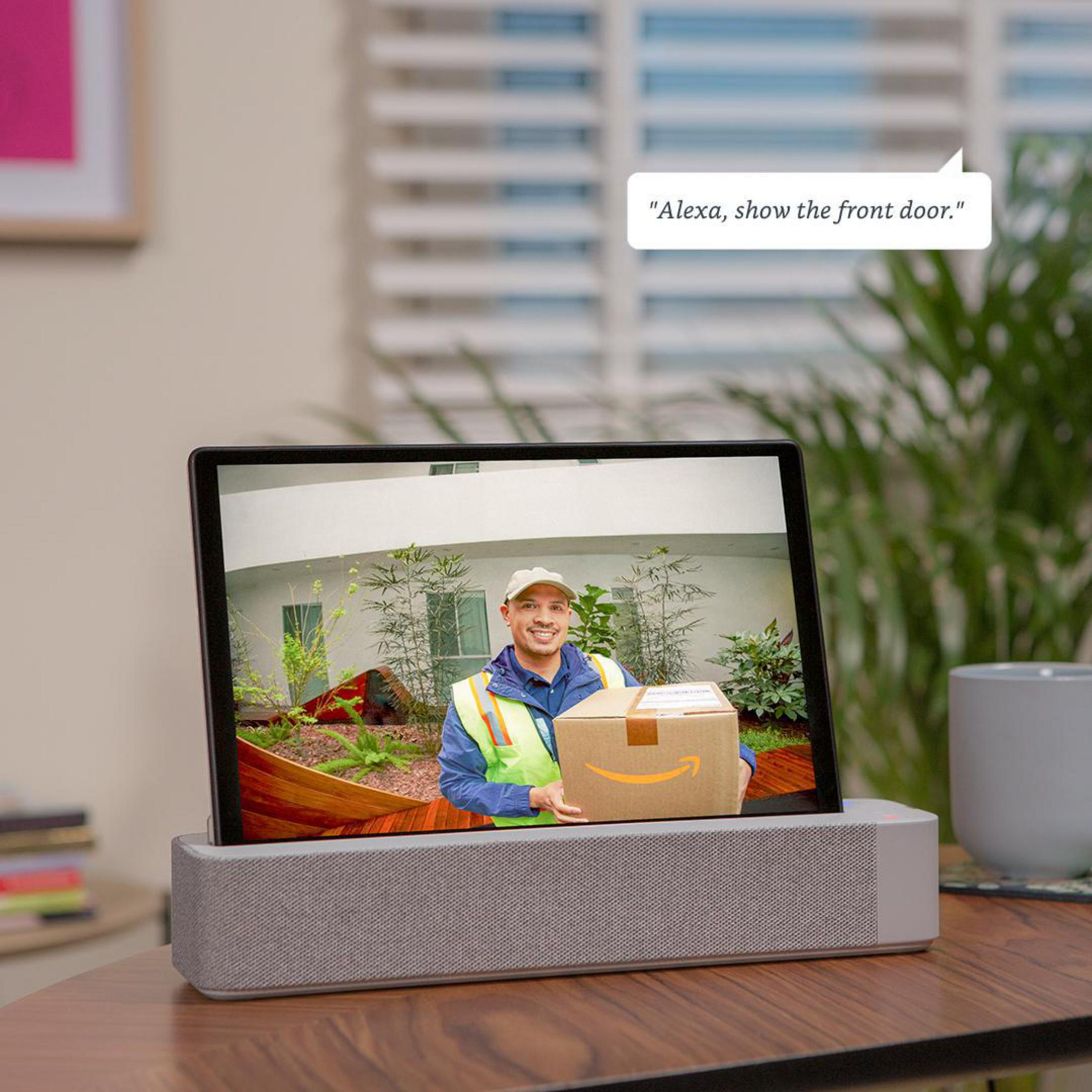 Tablet zeigt einen Zusteller. Textblase: 'Alexa, zeig die Haustür.' Graue Lautsprecherbasis.