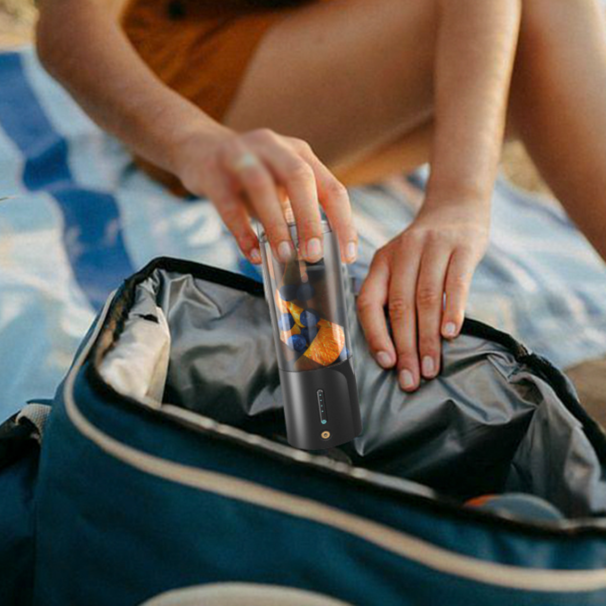 Eine Person legt einen Mixer mit Obst in eine blaue Tasche. Die Tasche ist am Strand.